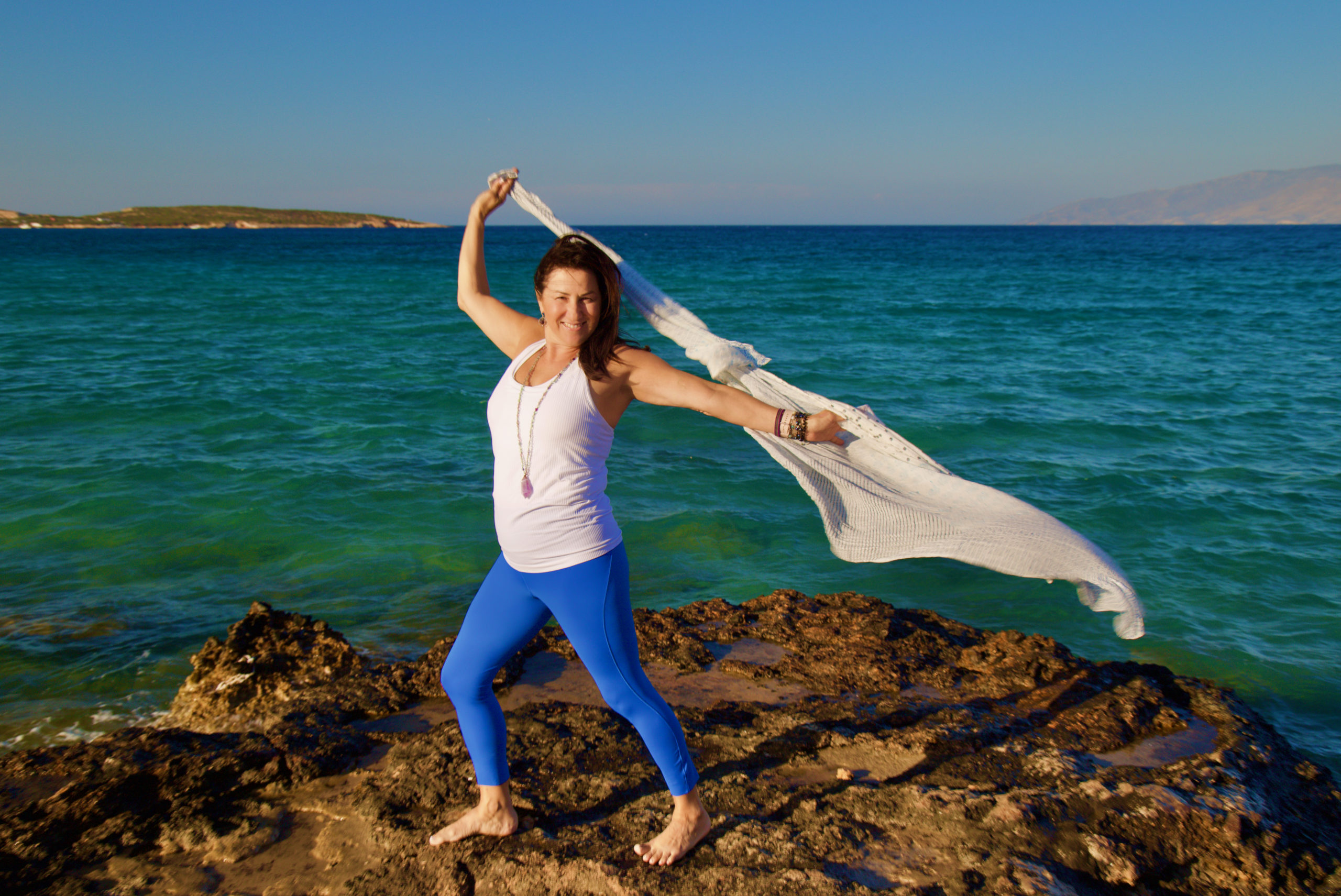 Yoga Student in Greece poses by the sea with billowing shawl in the wind behind her