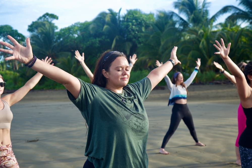 Student standing in Starfish Pose on the sand at sunrise during beach yoga practice.