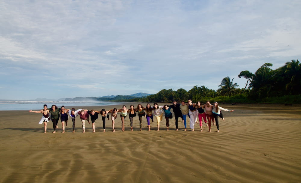 Yoga teacher training graduates in a line practicing Warrior III pose on the beach in Costa Rica.
